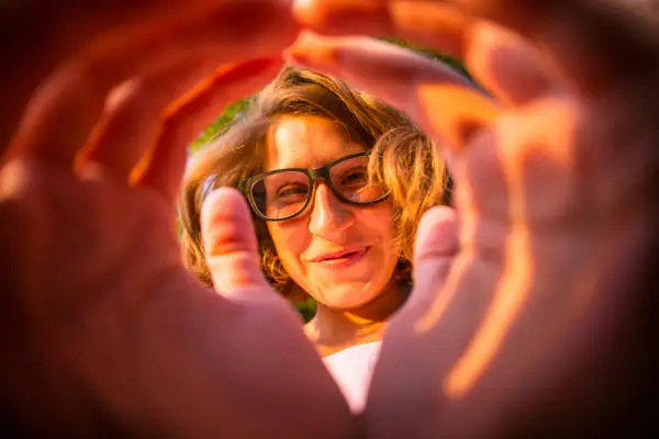 Ragazza con occhiali, sorride attraverso le mani che creano una cornice. Un'esperienza emozionale correttiva che illumina il volto.